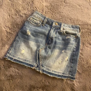 Blå lågmidja jeanskjol med slitningar - Säljer en blå jeanskjol med rå kant och slitna detaljer framtill. Kjolen har klassiska fickor både fram och bak samt knapp och dragkedja i midjan. Perfekt för en avslappnad och trendig look på en varm dag.