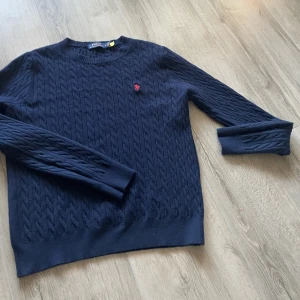 Mörkblå stickad tröja från Polo Ralph Lauren - Säljer en mörkblå stickad tröja från Polo Ralph Lauren med klassiskt kabelmönster och röd broderad logga på bröstet. Tröjan har rund hals och långa ärmar, perfekt för dig som gillar stilrena och tidlösa plagg.