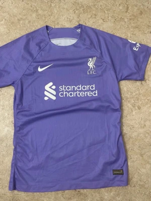 Lila Liverpool FC Nike matchtröja - Säljer en lila Liverpool FC matchtröja från Nike med korta ärmar och vit logga på bröstet. Tröjan har tryck med Standard Chartered och LFC-märket framtill. Perfekt för dig som älskar fotboll och vill visa ditt stöd för Liverpool.