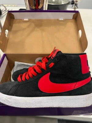 Nike SB Blazer Mid svart/röd - Nike SB Blazer Mid sneakers i svart mocka med röda detaljer och röd swoosh. Skorna har högt skaft, röda snören och vit platt sula med struktur. Insidan är vadderad för extra komfort och bakstycket har stor Nike-logga i svart. Endast använd en gång och kommer i orginalförpackning tillsammans med extra svarta snören.