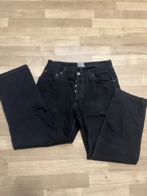 Svarta jeans från Henry Choice - Svarta jeans från Henry Choice med kontrastsömmar och klassisk femficksdesign. Jeansen har raka ben och normal passform, samt detaljerade nitar och logotyp på bakfickan. Materialet är robust bomull och de har knappgylf framtill.