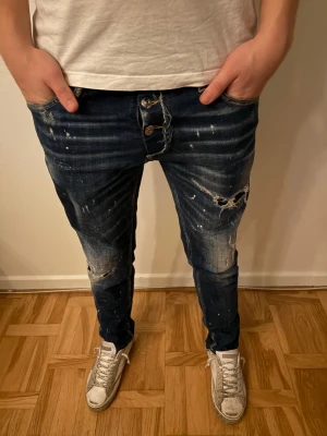 Dsquared2 Jeans - Säljer ett par blå jeans från Dsquared2 med skinny/slim passform. Jeansen har coola slitningar, färgstänk och trasiga detaljer på benen. Klassisk femficksmodell med knappgylf och Dsquared2-lapp bak i midjan. Perfekt för dig som gillar streetstyle och vill sticka ut. Kvitto finns!!!!