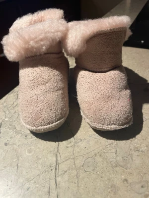 Beige Shepherd baby boots i fårskinn - Mysiga baby boots från Shepherd i ljusbeige färg med fluffig fårskinnskant. Utsidan är av mjuk mocka och insidan är fodrad med äkta fårskinn för extra värme. Rund tå och platt sula, perfekt för kalla dagar. Märke broderat på hälen.