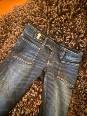 Blå bootcut jeans från Gina Tricot - Snygga blå jeans från Gina Tricot i storlek 158. Modellen har bootcut-ben, låg midja och klassiska fickor fram. Jeansen har två silverfärgade knappar och är gjorda i mjukt denim-material med lite stretch. Perfekta för dig som gillar retrovibbar och vill ha en cool siluett.