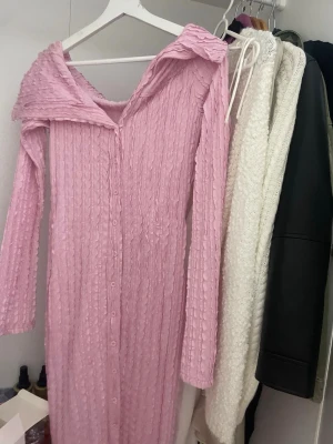 Lång rosa klänning med volangkrage - Säljer en lång rosa klänning med volangkrage och knappar hela vägen framtill. Klänningen har ett vågigt, strukturerat tyg och långa ärmar. Perfekt för dig som gillar unika detaljer och mjuka färger.