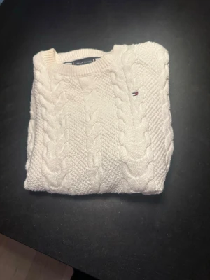 Vit stickad tröja Tommy Hilfiger - Säljer en vit stickad tröja från Tommy Hilfiger med klassiskt kabelstickat mönster och rund hals. Tröjan har en liten broderad logga på bröstet och är perfekt för kyliga dagar. Passar dig som gillar stilrena och tidlösa plagg.