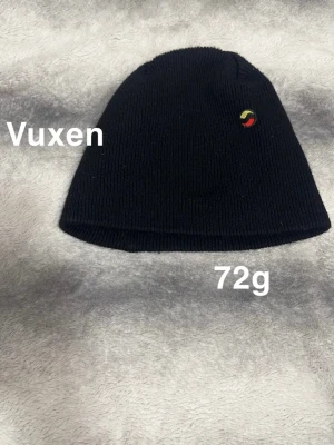 Svart stickad mössa med liten logga - Svart stickad mössa med en liten rund logga i rött, gult och grönt på sidan. Enkel och stilren design som passar till det mesta.🌸 Några noppor. Storlek vuxen