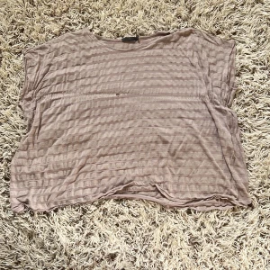 Beige randig topp från frk - Luftig och tunn topp från frk i beige med diskreta ränder. Modellen är kortärmad och har en avslappnad passform. Perfekt för dig som gillar basic med en twist. Materialet känns mjukt och lätt mot huden. Den har tyvärr några hål och det värkar som att någon har klippt av armarna. Storlek M