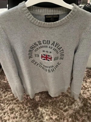 Grå stickad tröja Morris  - Snygg grå stickad tröja från Morris Gents i storlek XS. Tröjan har rund hals, ribbade muddar och tryck framtill med brittisk flagga och text. Perfekt för dig som gillar klassisk och stilren design.