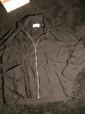 Svart overshirt från Ami - Säljer en svart overshirt från Ami Alexandre Mattiussi. Jackan har en clean look med dragkedja framtill, bröstficka och långa ärmar med knapp vid mudden. Tillverkad i bomull för en skön och lätt känsla. Perfekt att slänga över en hoodie eller t-shirt.