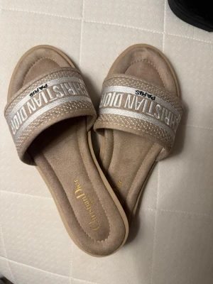 Beige sandaler  - Säljer ett par snygga beige sandaler med bred rem i mocka och broderad logga i vitt och silver. Mjukt innermaterial och platt sula för skön känsla. Perfekta till sommaren och riktigt trendiga att glida runt i. Aldrig använda💕passar 37 också