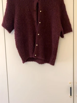 Vinröd kofta från Gina Tricot - Mysig vinröd kofta från Gina Tricot med korta ärmar och fluffig, stickad känsla. Koftan har dekorativa knappar framtill och en loose passform som ger en avslappnad vibe. Perfekt för lager på lager under höst och vinter.