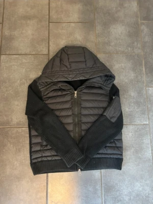 Svart Moncler pufferjacka med huva - Säljer en svart pufferjacka från Moncler med stickade ärmar och quiltad kropp. Jackan har huva, dragkedja framtill och en ficka med dragkedja på ena ärmen. Klassisk Moncler-logga på ärmen och ribbade muddar för extra komfort.