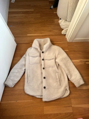 Beige teddyjacka med knappar - Mysig beige teddyjacka med stora bruna knappar framtill och två bröstfickor. Jackan har hög krage och långa ärmar, perfekt för kyliga dagar. Fodrad insida och avslappnad passform för en chill vibe.