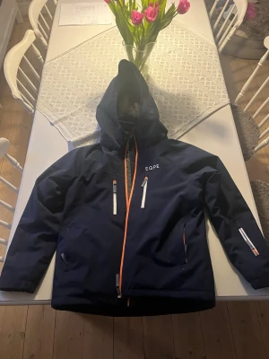 Mörkblå EQPE vindjacka med huva - Säljer en mörkblå skidjacka från EQPE med huva och orange dragkedja. Jackan har flera praktiska fickor med vita detaljer och en patch på ena ärmen. Perfekt för kyliga dagar i skidbacken. 