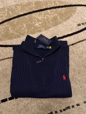 Marinblå stickad tröja Polo Ralph Lauren - Säljer en marinblå stickad tröja från Polo Ralph Lauren med ribbad struktur och röd broderad logga på bröstet. Tröjan har lång ärm, hög krage och half zip-dragkedja framtill. Perfekt för dig som vill ha en klassisk och stilren look.