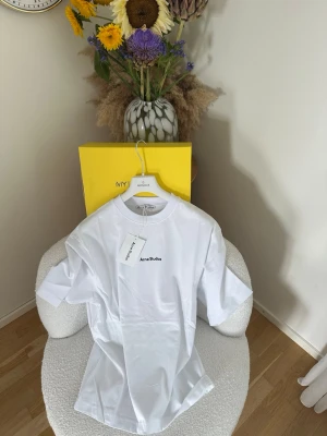 Vit t-shirt från Acne Studios - Clean vit t-shirt från Acne Studios med svart broderad logga på bröstet. Klassisk rund hals och rak passform. Tillverkad i 100% bomull för en skön och luftig känsla. Perfekt till jeans eller shorts för en enkel men snygg look.