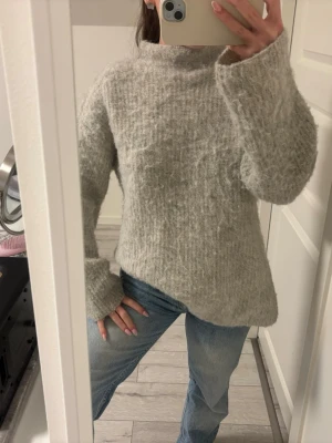 Ljusgrå stickad tröja med hög hals - Mysig ljusgrå stickad tröja med hög hals och lång ärm. Tröjan har en fluffig struktur och en relaxed, lite loose passform som ger en chill vibe. Perfekt att slänga på sig när det är kallt ute.