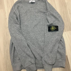 Grå sweatshirt från Stone Island - Säljer en grå sweatshirt från Stone Island med den klassiska loggan på vänster ärm. Tröjan har rund halsringning, ribbade muddar och är tillverkad i mjuk bomull. Perfekt för dig som gillar clean och stilren streetwear. Det är storlek s men den sitter lite baggy så den passar nog bäst som storlek M.