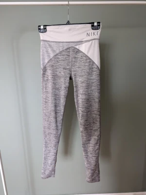 Gråmelerade leggings från NIKI - Supersnygga gråmelerade leggings från NIKI med hög midja och bred linning. De har en cool tvåfärgad design med ljusare grå upptill och mörkare grå nedtill. Perfekta för träning eller chill, i mjukt och stretchigt syntetmaterial.