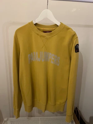 Gul tröja från Parajumpers - Gul tröja från Parajumpers med stor logga framtill och patch på ärmen. Klassisk rund halsringning och ribbade muddar. Perfekt för dig som vill ha en clean men ändå statement look.