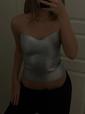 Silver axelbandslös topp från Zara - Säljer en metallic silver axelbandslös topp från Zara i storlek S. Toppen har en snygg bandeauform med lätt glansigt material och en figurnära passform som ger en modern look. Perfekt till dig som vill  ha till någon fest !.