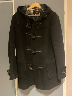 Svart duffel från Gloverall - Klassisk svart duffeljacka från Gloverall med huva och träknappar. Jackan har stora fickor framtill och rutigt innerfoder i grönt, blått och gult. Tillverkad i ull och perfekt för kalla dagar. Snygg och tidlös brittisk stil.                                                          Mått: Ärm-64cm Armhåla och nedåt-50cm Krage och nedåt-71cm Nacke och nedåt-80cm PRIS KAN DISKUTERAS💕