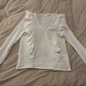 Vit blus med volang från Vila - Säljer en superfin vit blus från Vila med v-ringning och långa ärmar. Blusen har en strukturerad, lite bubblig yta och dekorativa volanger som går från axlarna ner längs framsidan. Perfekt för dig som gillar romantiska detaljer och en feminin look.