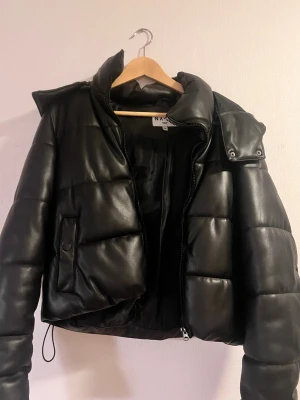 Svart pufferjacka i skinnimitation NA-KD - Säljer en svart pufferjacka från NA-KD i skinnimitation. Jackan har hög krage, avtagbar huva och dragkedja framtill. Den är quiltad och har snörning i nederkant för en snygg passform. Perfekt för kalla dagar och riktigt trendig look. Har ett litet märke vid luva se bild snart är det inget som syns mycket. Mycket snygg jacka. 