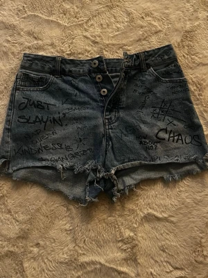 Blå jeansshorts med graffiti-tryck - Unika blå jeansshorts med handskrivna graffiti-texter och råa, fransiga kanter. Shortsen har hög midja, tre knappar framtill och klassiska fickor både fram och bak. Perfekta för dig som vill sticka ut med en edgy och personlig stil.