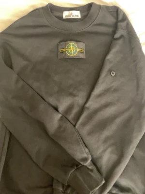 Svart sweatshirt från Stone Island - Säljer en svart sweatshirt från Stone Island med den ikoniska kompasspatchen på ärmen. Tröjan har rund hals, ribbade muddar och är tillverkad i mjuk bomull. Perfekt för dig som gillar streetwear och vill ha en clean look.