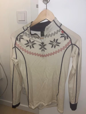 Beige halvzip tröja Helly Hansen - Säljer en beige långärmad tröja från Helly Hansen med snyggt vintermönster i svart och rött över bröstet. Tröjan har half zip och mörka kontrastsömmar samt muddar. Perfekt för kalla dagar och outdoor-aktiviteter.