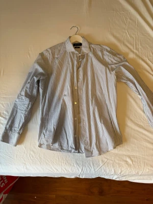 Randig skjorta från Riley slim fit - Säljer en stilren vit skjorta med smala svarta ränder från Riley. Skjortan är slim fit och har klassisk krage, långa ärmar och knäppning framtill. Perfekt för dig som gillar en clean och snygg look.