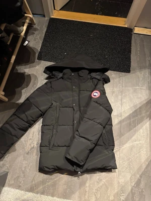 Canada goose jacka  - Säljer en svart dunjacka från Canada Goose i storlek S. Jackan har en avtagbar huva med fet pälskant, stora fickor och klassisk patch på ärmen. Perfekt för kalla dagar och riktigt snygg till vinterlooken.