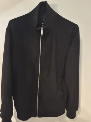  Zara Jacka - Säljer en svart bomberjacka från Zara med hög krage och hel dragkedja framtill. Jackan har en clean look utan synliga fickor eller mönster och är tillverkad i ett mjukt material som känns skönt mot huden. Perfekt för dig som gillar minimalistisk stil. Storlek S 