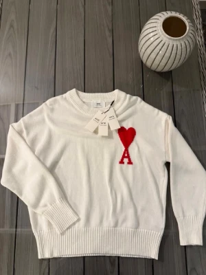 Vit stickad tröja från Ami Paris - Säljer en vit stickad tröja från Ami Paris med röd hjärtlogga och bokstaven A på bröstet. Tröjan har rund hals, ribbade muddar och är långärmad. Perfekt för dig som gillar stilrena plagg med en twist.