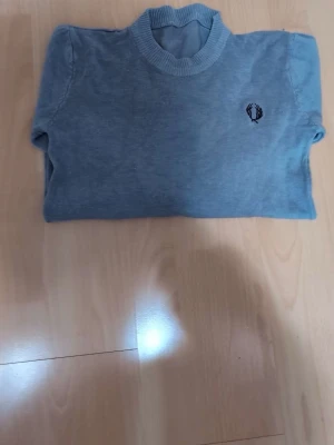 Blå stickad tröja från Fred Perry - Säljer en klassisk blå stickad tröja från Fred Perry med rund halsringning och den ikoniska lagerkransen broderad på bröstet. Tröjan har ribbade muddar vid ärmslut och nederkant samt lång ärm. Perfekt för dig som gillar stilren och enkel design.