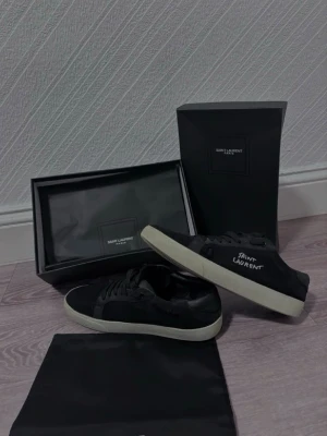 Svarta Saint Laurent sneakers - Säljer ett par svarta Saint Laurent sneakers med vit sula och handskriven logga på sidan. Skorna har snörning och är tillverkade i mocka och skinn, vilket ger en lyxig känsla. Kommer med originalkartong och dustbag. Lite andvända 