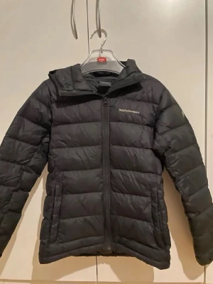 Svart pufferjacka Peak Performance - Säljer en svart pufferjacka från Peak Performance med huva och dragkedja framtill. Jackan har en klassisk quiltad design och är perfekt för kyligare dagar. Långärmad modell med en clean look och diskret logga på bröstet.