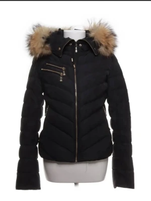 Svart dunjacka från Hollies! - Säljer en svart pufferjacka från Hollies med stor fluffig huva, jackan har även guldiga dragkedjor Perfekt för kalla dagar. Storlek 38 säljer då jag inte behöver den längre!