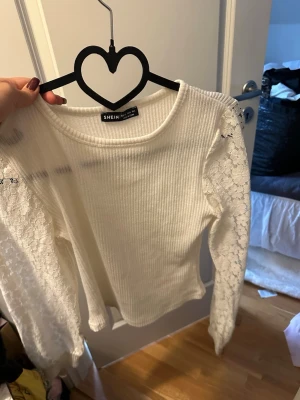 Vit ribbad topp med spetsärmar från SHEIN - Säljer en vit ribbad topp från SHEIN i storlek XS. Toppen har långärmade spetsärmar med blommigt mönster och en rund halsringning. Kroppen är i stretchigt ribbat material och ärmarna är genomskinliga med fina spetsdetaljer.