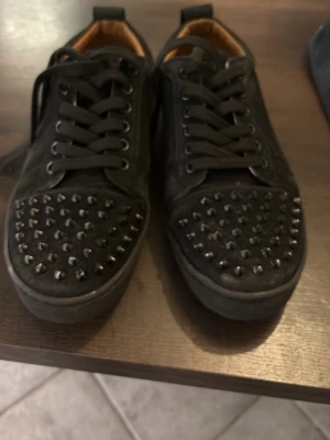 Svarta sneakers med nitar från Louboutin - Säljer ett par svarta sneakers från Louboutin med klassisk röd sula och coola svarta nitar på tån. Skorna har snörning och är tillverkade i mocka för en lyxig känsla. Perfekta för dig som vill sticka ut med en edgy stil.