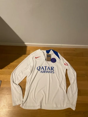 PSG X Nike Tracksuit 25/26 Player Version Helt Ny!!! - Säljer ett helt nytt träningsset från Paris Saint-Germain, säsongen 2025/2026. Setet består av en långärmad vit träningströja och svarta träningsbyxor – båda med PSG:s officiella klubbmärke och Nike-logga i rött. Tröjan har quarter-zip och tryckt sponsor “Qatar Airways” på bröstet. Byxorna har elastisk midja med snörning, blå sidodetalj och matchande emblem.                                                                                 Helt Ny!!!