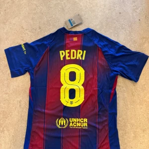 FC Barcelona Pedri matchtröja Nike - Snygg FC Barcelona matchtröja från Nike med Pedri och nummer 8 på ryggen. Tröjan har klassiska blå och röda ränder, gula detaljer och klubbmärke på bröstet. Tillverkad i lätt och ventilerande material, perfekt för fotboll eller att bära som supporter.