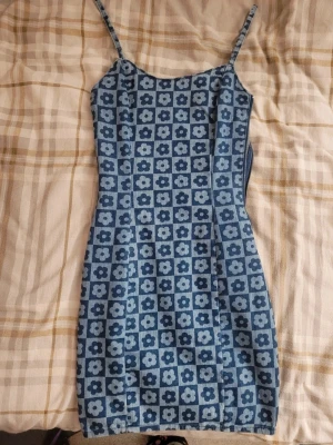 Blå blommig kortklänning med smala band - Supersöt blå kortklänning med smala axelband och ett retro blommigt mönster i olika blå nyanser. Klänningen har figurnära passform och dragkedja i sidan. Perfekt för dig som gillar vintagevibbar och vill sticka ut.