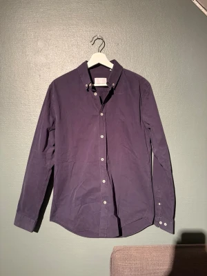 Mörkblå skjorta från Samsøe Samsøe - Snygg mörklila skjorta från Samsøe Samsøe i storlek M. Klassisk modell med knappar hela vägen, button down-krage och långa ärmar. Skjortan är tillverkad i bomull och har en clean look utan mönster eller extra detaljer.
