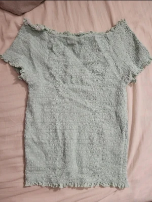 Ljusgrön ribbad topp med volang - Säljer en ljusgrön ribbad topp med korta ärmar och volangkant vid hals, ärmslut och nederkant. Toppen har en stretchig, smockad struktur som ger en tight passform och en söt, feminin look. Perfekt till jeans eller kjol!