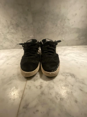 Svarta och vita sneakers med snörning - Säljer ett par svarta och vita sneakers med klassisk låg profil och snörning. Skorna har svart ovandel i mocka och detaljer i vitt skinn på sidor och häl. Sulan är vit och platt, och tån är rundad. Perfekta för dig som gillar en enkel men stilren look.