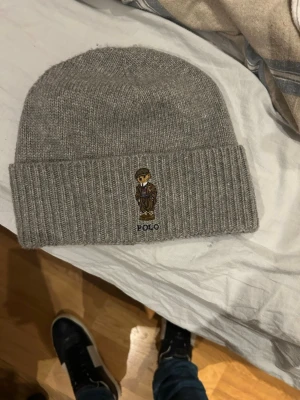  Polo Ralph Lauren mössa - Säljer en stilren grå mössa från Polo Ralph Lauren med broderad nalle på framsidan. Mössan är ribbstickad och tillverkad i ullblandning och återvunnen polyester. Perfekt accessoar för kyliga dagar.