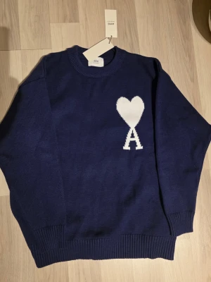 Marinblå stickad tröja från Ami - Säljer en marinblå stickad tröja från Ami med ikoniskt vitt hjärta och A på bröstet. Tröjan har rund halsringning, ribbade muddar och är långärmad. Perfekt för dig som gillar stilrena detaljer och vill ha något som sticker ut.